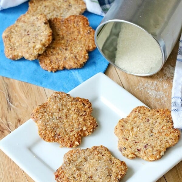 Four homemade hazelnut cookies rest on a white plate, with more cookies and a sugar canister in the background.