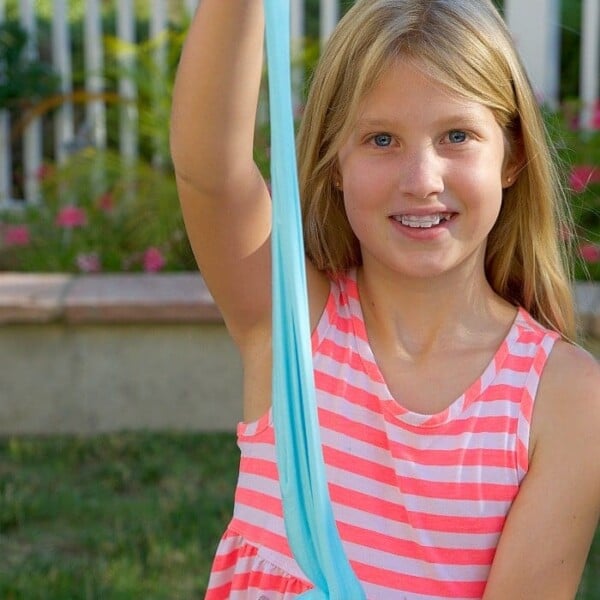 In a garden, a child wearing a pink striped dress stretches a long length of blue slime. The scene sparks curiosity about how to make fluffy slime just like it.