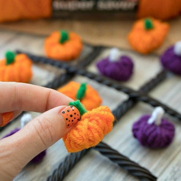 Hand holding a crocheted orange pumpkin for a yarn pumpkin craft tic-tac-toe game, displayed on a wooden surface.