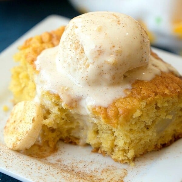 A slice of banana cake topped with a scoop of vanilla ice cream rests elegantly on a white square plate, evoking the comforting flavors of banana pudding.