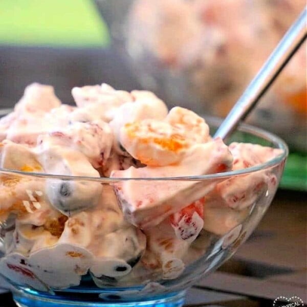 A bowl of creamy fruit salad with a spoon, featuring grapes, oranges, marshmallows, and a dollop of cool whip.