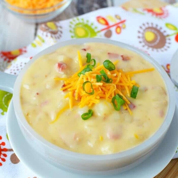 Crockpot loaded baked potato soup, creamy and delicious, is topped with shredded cheese and green onions. It's elegantly presented in a white bowl on a patterned cloth.