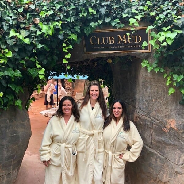 Three people in robes smile under a Club Mud sign at Glen Ivy Spa, Corona, surrounded by lush greenery and a rocky path.