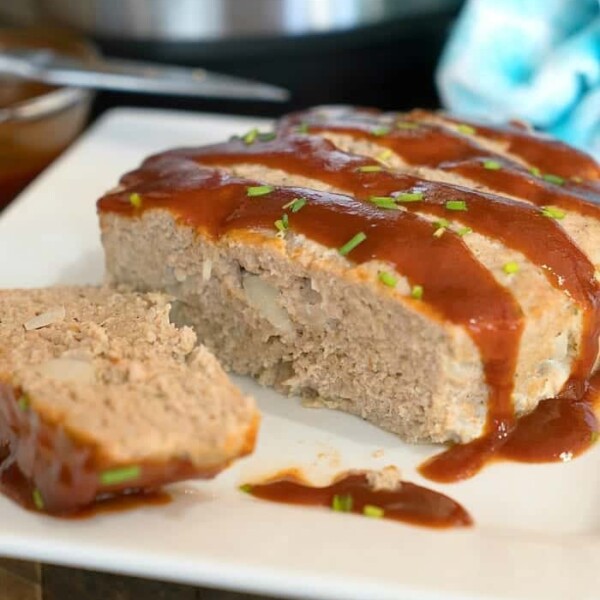 A sliced turkey meatloaf topped with a glossy brown sauce and garnished with chopped green onions sits on a white square plate. The Instant Pot, which made this tender loaf possible, is visible in the blurred background alongside a blue cloth.