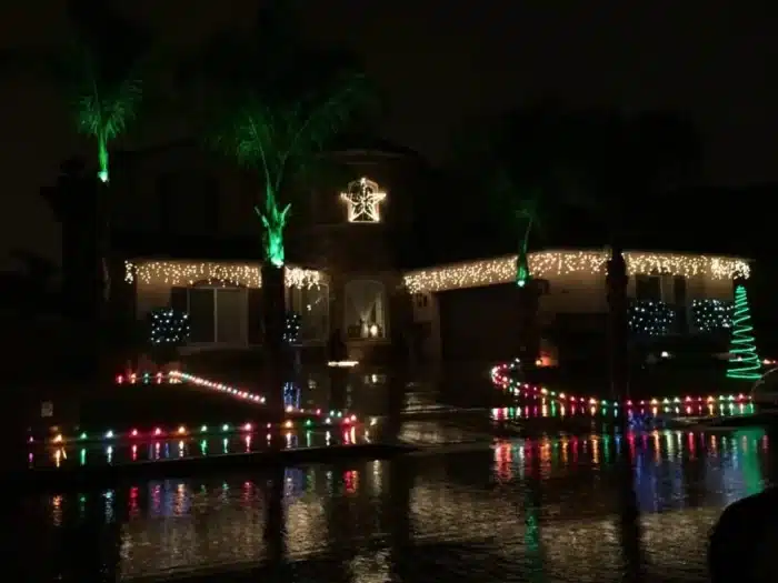 A house in Temecula is decorated with colorful Christmas lights. Green lights illuminate the palm trees, while white and multicolored lights adorn the house and yard. A star decoration is displayed above the entrance, and wet pavement reflects the festive glow at night.