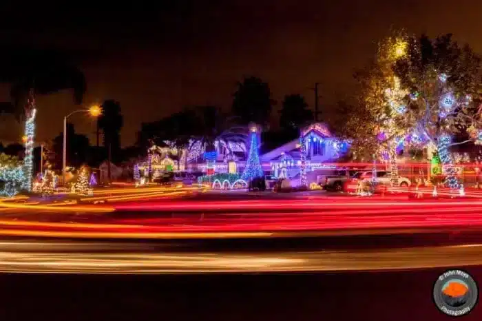 A festive neighborhood in Temecula at night is illuminated with colorful Christmas lights, displaying various decorations. Light trails from passing vehicles create streaks across the foreground, adding dynamic movement to the scene, like a lively map of holiday cheer.