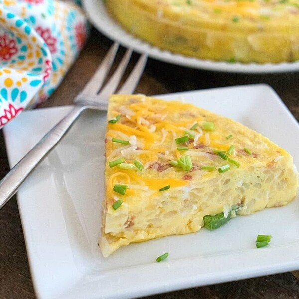 Slice of frittata garnished with chives and cheese on a square white plate, with a fork beside it. A full instant pot breakfast casserole graces a round plate in the background, next to a colorful cloth.