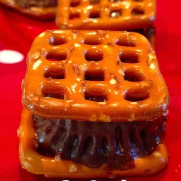 A piece of chocolate candy is nestled between two square pretzels, reminiscent of Reese's Pretzel Bites, resting on a red polka dot surface.