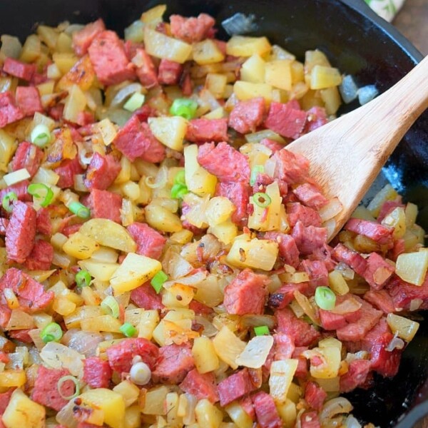 A skillet containing a homemade corned beef hash recipe with diced corned beef, potatoes, onions, and green onions is being stirred with a wooden spoon.