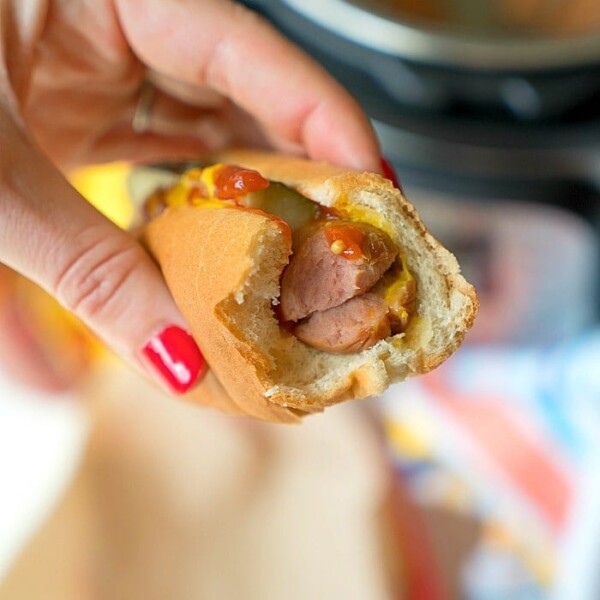 A hand holds a bitten hot dog with mustard and ketchup, the vibrant red nails adding a pop of color. Perfectly cooked using an Instant Pot, this hot dog promises both convenience and flavor in every bite.