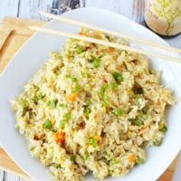 A plate of pressure cooker fried rice with peas and carrots rests on a wooden board, accompanied by chopsticks and a bottle of soy sauce.