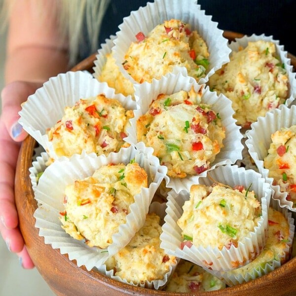 A person holds a wooden bowl filled with Bisquick savory breakfast muffins, topped with herbs and diced peppers in paper liners.