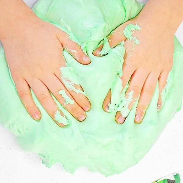 Hands kneading light green slime made with shaving cream on a white surface, with part of a green-patterned cloth visible nearby.