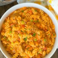 A bowl of rotini pasta with creamy tomato sauce, bell peppers, and chives sits beside an Instant Pot and a floral napkin, reminiscent of a cheesy goulash twist.