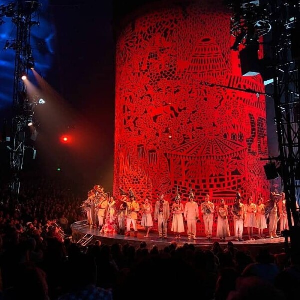 A theatrical performance reminiscent of Cirque du Soleil, with a cast on stage, an intricate red backdrop, and an audience in a dimly lit venue, invites attendees into the mesmerizing world of Luzia.