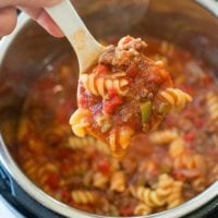 A hand holds a white ladle filled with Instant Pot sloppy joe casserole—rotini pasta, ground meat, and rich tomato sauce—over an open pressure cooker brimming with the same delicious mixture.