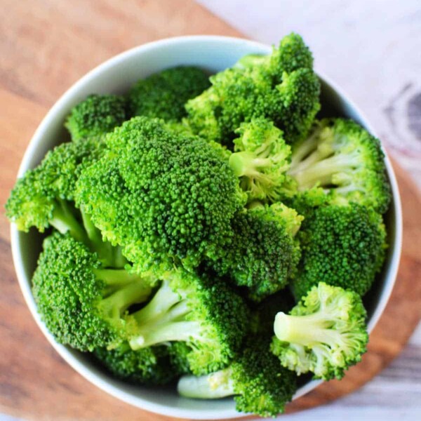 A bowl filled with fresh green broccoli florets, perfect for a delicious steamed broccoli recipe, is placed on a wooden cutting board. The background shows a lightly textured white surface.