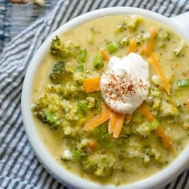 Bowl of quick broccoli cheese soup topped with shredded carrots, green onions, and a dollop of sour cream on a striped cloth.