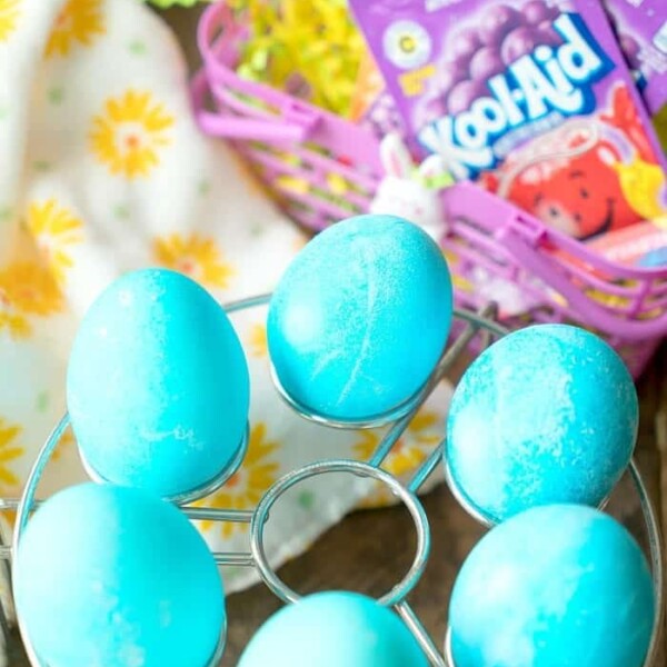 Blue dyed eggs rest elegantly on a metal stand, while a basket brimming with Kool-Aid packets and decorative yellow grass sits in the background. Discover how to dye eggs with Kool-Aid for vibrant hues that brighten any table setting.