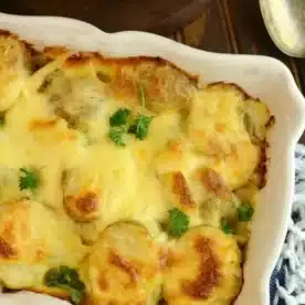 A baking dish with easy potatoes au gratin, topped with a cheesy crust and garnished with parsley, sits on a wooden table alongside a serving spoon and plates.