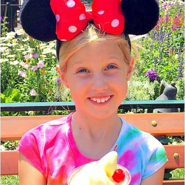 A young girl with blonde hair wearing Minnie Mouse ears and a tie-dye shirt beams as she holds a cup of Dole Whip with a cherry on top. Sitting on a bench in a garden-like setting, she embodies the joy of discovering how to eat for free at Disneyland.