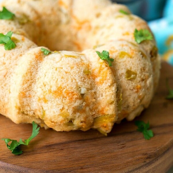 A bundt-shaped savory beer bread rests on a wooden board. The bread is lightly browned with small visible chunks of cheese and herbs. Fresh parsley leaves are scattered on top for garnish, offering a delightful twist to the classic instant pot recipe.