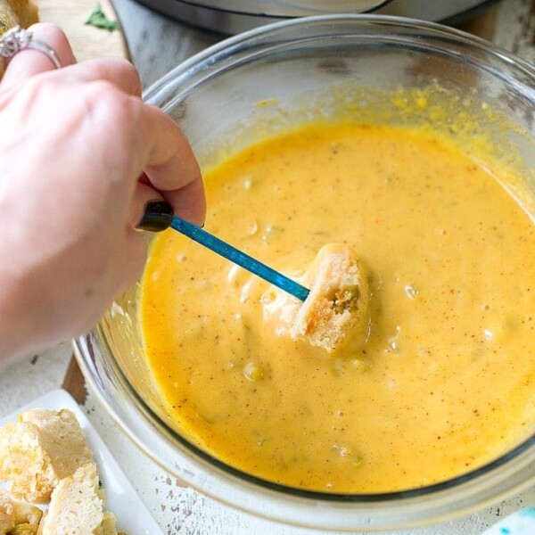 A bowl of yellow cheese fondue with broccoli invites a hand to dip a piece of bread using a blue fork. An Instant Pot pressure cooker sits in the background, while a plate with bread chunks waits on the side.