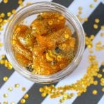 A jar of golden, glittery gummy bears that look like honey slime, with sequins on a black and white striped background.