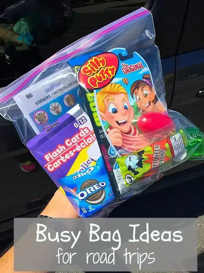 A hand holds a clear ziplock bag brimming with road trip busy bag essentials like Silly Putty, flashcards, Oreo cookies, and a small juice pouch. The text on the image reads, "Busy Bag Ideas for road trips.