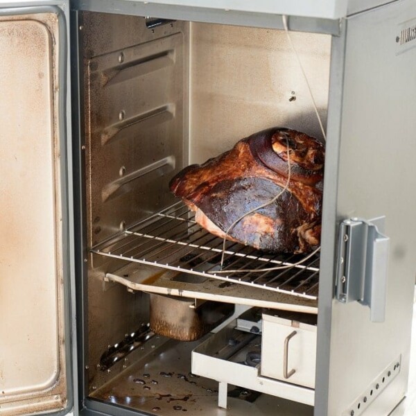 A metal smoker with the door open reveals a large smoked cooked ham on the rack inside. The smoker rests on a wooden surface.