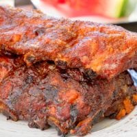 A plate with two racks of seasoned, grilled ribs showcases the best way to smoke ribs. In the background, there's a slice of watermelon on a table.