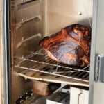 A cooked ham rests on a rack inside the smoker, tendrils of smoke curling around it. Text above reads, "Smoking a Cooked Ham." The smoker's door is open, showcasing the perfect setup for infusing delicious flavors.