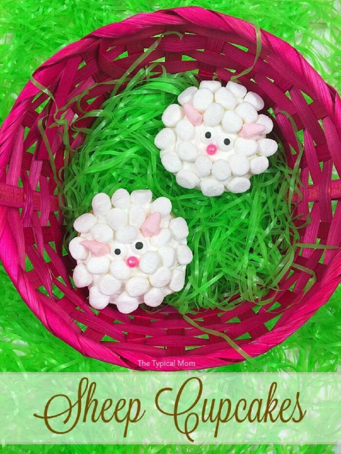 A bright pink basket with green grass-like filler holds two cute cupcakes decorated as sheep, using white marshmallows for the wool, pink marshmallows for ears, googly eyes, and a pink candy for noses. The text on the image reads "Sheep Cupcakes.