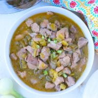A bowl of hearty chili verde, filled with tender chunks of meat and green onions, garnished with vibrant chopped vegetables. The dish sits on a white surface next to fresh green onions, with a colorful patterned cloth and the gleaming lid of a pressure cooker in the background.