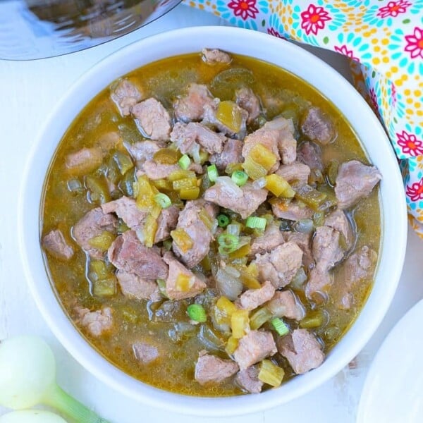 A bowl of hearty chili verde, filled with tender chunks of meat and green onions, garnished with vibrant chopped vegetables. The dish sits on a white surface next to fresh green onions, with a colorful patterned cloth and the gleaming lid of a pressure cooker in the background.