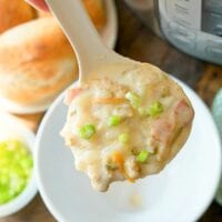 A ladle of creamy, easy Instant Pot clam chowder with diced vegetables is topped with chopped green onions. It's held above a white bowl, while an Instant Pot sits in the background alongside rolls and a small bowl of green onions on the wooden table.