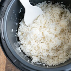 A Crockpot Express revealing a batch of perfectly cooked white rice. A white plastic serving spoon gracefully rests on the rice, ready for serving. Text at the bottom reads Crockpot Express White Rice, signifying simple perfection in every grain.