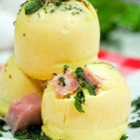 Three steamed egg bites, reminiscent of Starbucks' sous vide eggs, are stacked on a white plate, garnished with chopped parsley and small pieces of ham. A red and white checkered cloth serves as a backdrop.