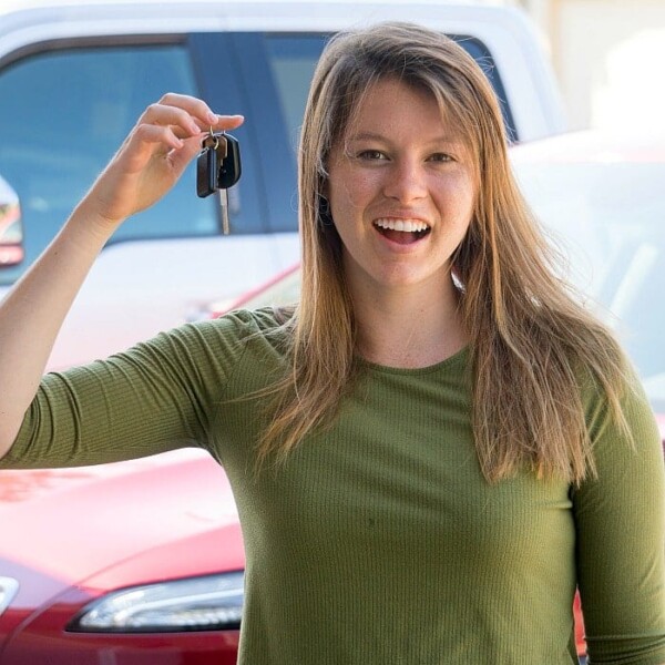 A woman in a green shirt smiles while holding car keys, possibly celebrating a teen driving contract. She stands outside in front of a red car and a white SUV in a residential area.