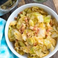 A bowl of pressure cooker fried cabbage with crispy bacon pieces sits on a wooden table, accompanied by a small bowl of freshly chopped chives.