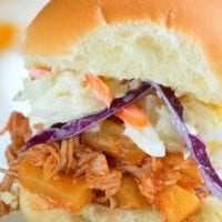 A close-up of an Instant Pot Hawaiian pulled pork sandwich, featuring tender shredded meat, pineapple pieces, and coleslaw inside a soft bun. The layers of colorful ingredients are visible, with vibrant purple cabbage and carrot strands enhancing the coleslaw.