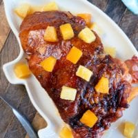 A glazed ham with pineapple chunks sits invitingly on a white platter, reminiscent of a delicious crockpot ham recipe, all elegantly arranged on a wooden table adorned with a blue napkin.