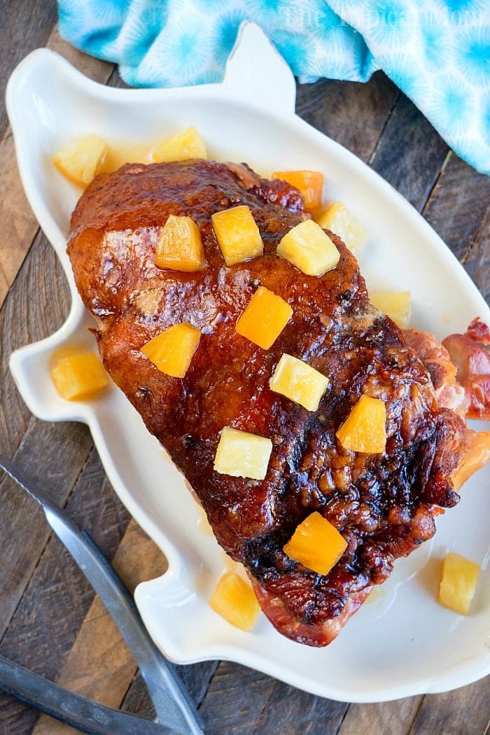 A glazed ham with pineapple chunks sits invitingly on a white platter, reminiscent of a delicious crockpot ham recipe, all elegantly arranged on a wooden table adorned with a blue napkin.