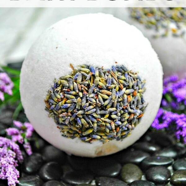 A large white handmade bath bomb with dried lavender rests on black stones, surrounded by purple flowers.