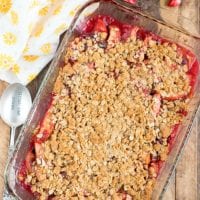 A glass baking dish filled with a cranberry apple cobbler, featuring sliced apples and cranberries topped with a golden oat crumb layer. It is placed on a wooden surface, surrounded by a spoon, a yellow-patterned cloth, an apple, and an orange slice.