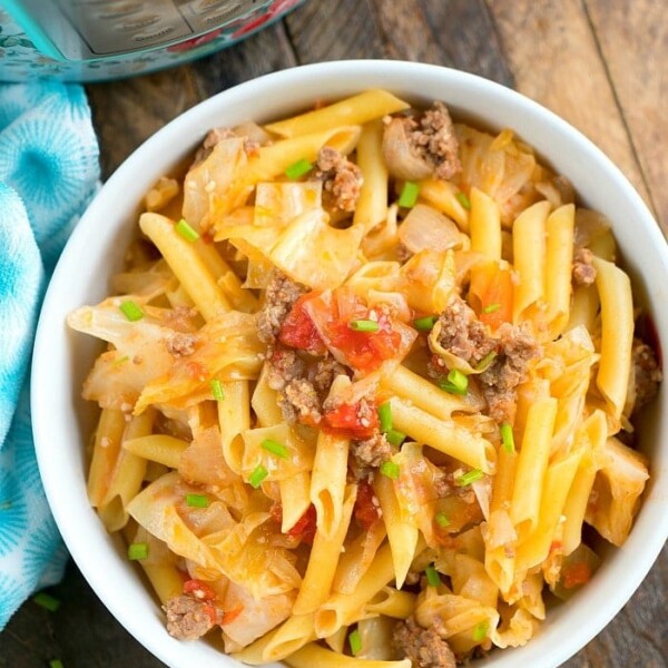 A bowl filled with penne pasta, perfectly cooked in a pressure cooker, mixed with ground beef, chopped tomatoes, and savory sausage. The dish is topped with green onions and accompanied by crisp cabbage on a wooden table beside a blue patterned cloth.