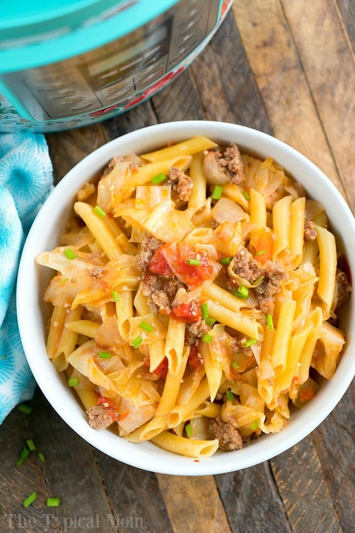 A bowl filled with penne pasta, perfectly cooked in a pressure cooker, mixed with ground beef, chopped tomatoes, and savory sausage. The dish is topped with green onions and accompanied by crisp cabbage on a wooden table beside a blue patterned cloth.