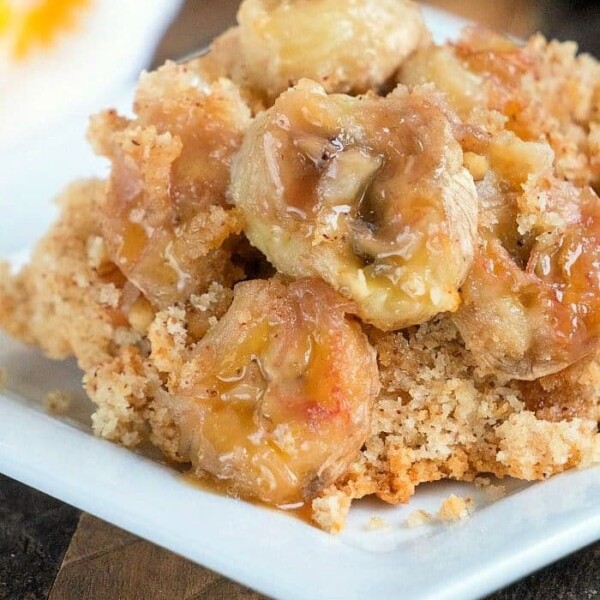 A close-up of a portion of banana dump cake on a white plate showcases slices of banana beneath a crispy, golden-brown topping. It rests on a wooden surface, with part of another dish visible in the background.