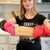 A girl in a black shirt proudly holds a loaf of whole wheat bread in a bag with red oven mitts, standing confidently in her kitchen.