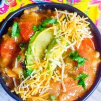 A bowl of Instant Pot turkey soup topped with sliced avocado, shredded cheese, and chopped cilantro. The bowl rests on a wooden surface next to a colorful, patterned fabric. The soup features chunks of vegetables in a rich tomato-based broth.